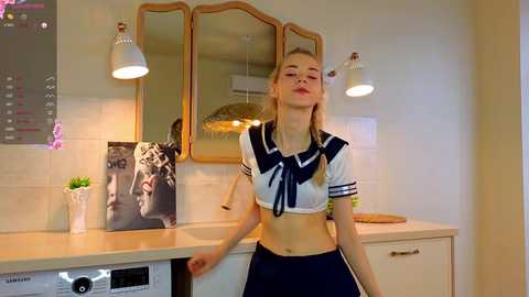 Media: A young, slender, blonde woman in a sailor-style crop top and high-waisted skirt poses confidently in a modern kitchen. The background features a tiled wall, a stove, and a decorative fish poster.