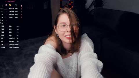 Media: Video of a young woman with glasses, light skin, and shoulder-length brown hair, wearing a white off-shoulder sweater, smiling and leaning forward on a dark couch.