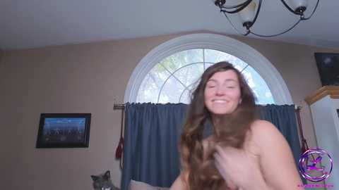 Media: Video of a smiling, nude, fair-skinned woman with long, wavy brown hair, holding her hair, standing in a bedroom with a large arched window, blue curtains, and a framed picture on the beige wall.