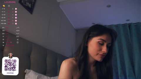 Media: Video of a young woman with long black hair, fair skin, and a slender physique, topless, sitting on a bed with a grey headboard. The room is dimly lit with teal curtains, and a \"LOUISIANA\" watermark appears in the top left corner.