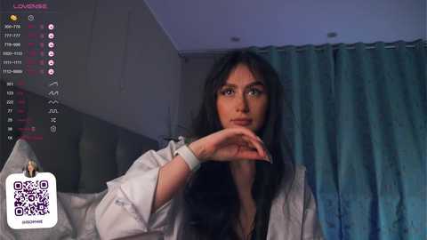 Media: A video of a young woman with long black hair, wearing a white lab coat, standing in a dimly lit room with teal curtains, looking contemplative.