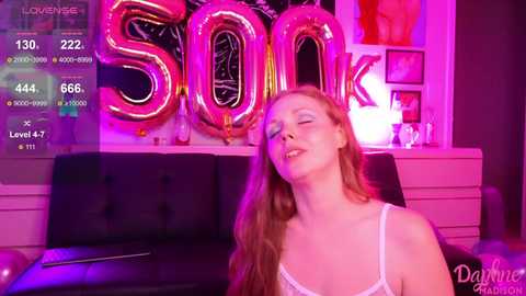 Media: A video of a fair-skinned, red-haired woman with long hair, wearing a white lace bra, lying back on a black couch, surrounded by large pink and red \"SOON\" balloons in a dimly lit room.