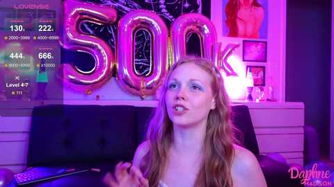 Media: Video of a blonde woman with long hair, wearing a strapless top, standing in a dimly lit room with purple and pink lighting. Large, shiny \"SOON\" letters and a digital timer display on a wall.