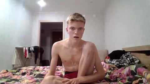 Media: Video of a shirtless, fair-skinned, young man with short blonde hair, sitting on a bed with a floral-patterned blanket in a sparsely furnished room.