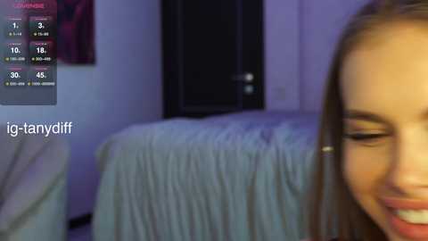 Media: A video of a smiling young woman with light skin and long brown hair, captured in a dimly lit bedroom with a white bedspread.