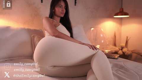 Media: Video of a curvaceous, fair-skinned woman with long black hair, wearing tight white leggings, sitting on a beige sofa in a dimly lit, cozy room.