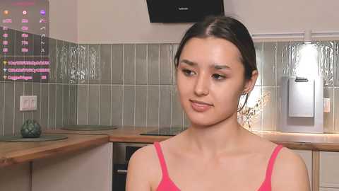 Media: Video of a young woman with dark hair in a pink tank top, standing in a modern kitchen with green tiled backsplash, a fridge, and wooden countertop.