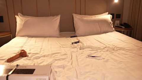 Media: Video of a hotel bed with white linens, two large white pillows, a folded sheet, and a digital alarm clock on the nightstand. The background features a beige wall and a wooden nightstand.