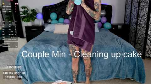 Media: Video of a tattooed woman in a blue dress, holding a purple towel, standing in a bedroom with a bed, balloons, and a TV screen displaying \"Balloon Puke 3: 8 weeks, 100 lbs.\