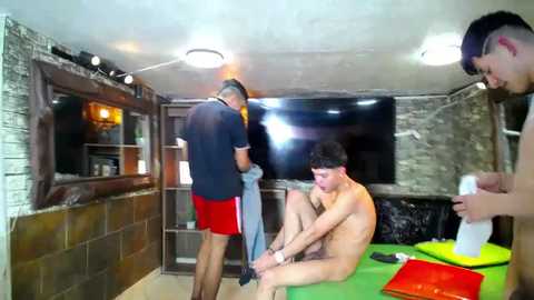 Media: Video of a dimly-lit, rustic indoor room with three men: one nude, one partially clothed, and one in a black cap and red shorts. They are sitting on a green table, with a stone wall and a window with curtains in the background.