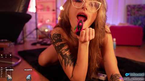 Media: Video of a topless, light-skinned woman with long blonde hair, glasses, and tattoos, licking a black dildo, in a dimly lit, messy room.