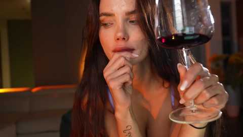 Media: Video of a brunette woman with long hair, wearing a sleeveless top, holding a wine glass, with a contemplative expression.