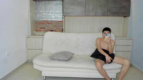 Media: Video of a young, shirtless Asian man with short dark hair, wearing black shorts, sitting on a white leather couch in a minimalist, white-walled room with a brick accent wall and metallic kitchen cabinets.