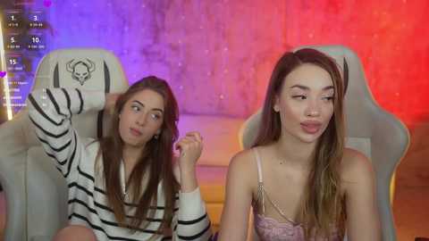 Media: Video of two young women with light skin, one in a striped sweater, the other in a lace top, seated in gaming chairs against a colorful LED-lit backdrop.