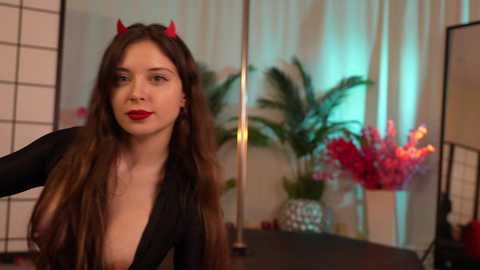 Media: Video of a woman with long brown hair, wearing red devil horns and a plunging black dress, standing in front of a floral arrangement and white folding screens.