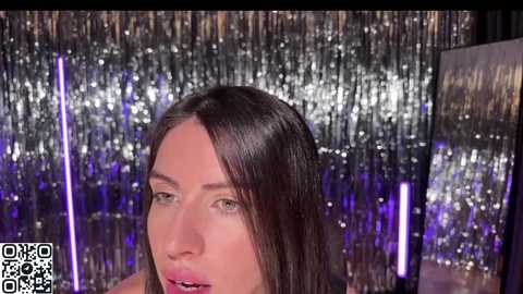 Media: Video of a woman with straight, dark hair, wearing makeup, in a dimly lit room with silver tinsel and purple lighting. QR code visible in the bottom left corner.