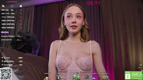 Media: Video of a fair-skinned woman with long, wavy blonde hair, wearing a pink floral bralette, standing in a dimly lit bedroom with a digital live stream overlay, showing viewers' chat messages.