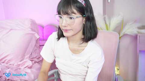 Media: Video of a young Asian woman with straight black hair, wearing glasses and a white top, sitting on a pink chair in a softly lit bedroom with pink and white decor.