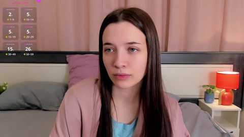 Media: Video of a young Caucasian woman with long dark hair, wearing a pink jacket, sitting on a bed with gray sheets and a red lamp, beige curtains in the background.