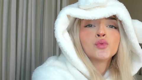 Media: Video of a young, fair-skinned, blonde woman with light makeup, wearing a fluffy white bear hood, puckering her lips. Background features a blurred curtain and neutral wall.