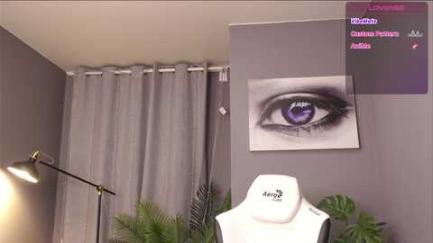 Media: Video of a modern bedroom with a large gray curtain, a purple-eyed artwork on a gray wall, a green fern, and a white Armani necklace on a white stand.