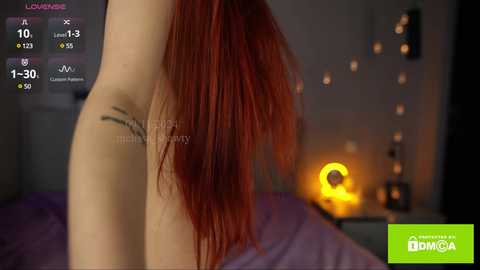 Media: Video of a woman with long, straight red hair, partially nude, showing her side profile and arm tattoo. Background features a dimly lit bedroom with a warm, yellow lamp and indistinct decor.