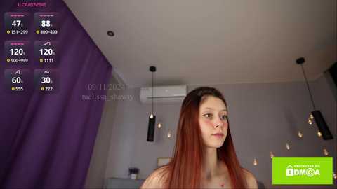 Media: Video of a young woman with long, straight auburn hair, wearing a red top, standing in a modern, minimalistic living room with gray walls, purple curtains, and hanging pendant lights.