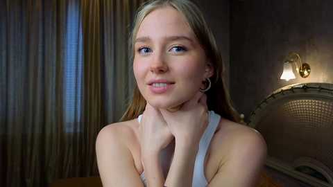 Media: Video of a young, fair-skinned woman with long blonde hair, wearing a white tank top, standing in a dimly lit room with beige curtains, a wicker headboard, and a wall-mounted lamp.