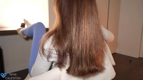 Media: Video of a person with long, straight brown hair, wearing a white shirt and blue leggings, sitting in a dimly lit room with a window and closed door in the background.