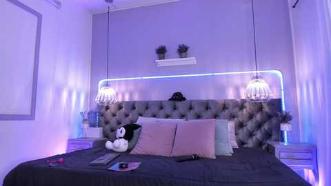 Media: Video of a modern bedroom with a tufted gray headboard, purple accent lighting, two hanging glass pendant lights, and plush pillows in pastel colors.