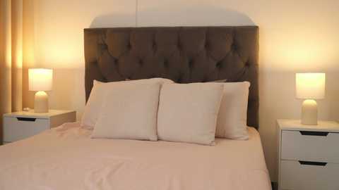 Media: Video of a modern bedroom with a plush, tufted dark gray headboard, two white pillows, beige sheets, and a white nightstand on each side, each holding a cylindrical lamp.