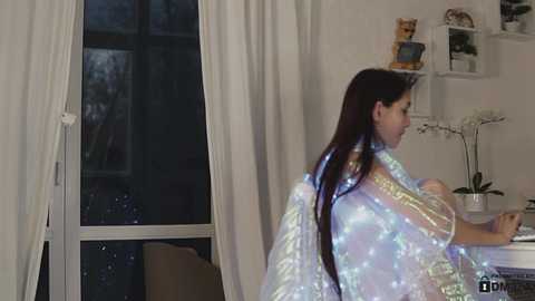 Media: Video of a woman with long, dark hair wearing a luminous, iridescent gown, standing in a modern, softly lit room with white walls, a window, and a potted orchid.