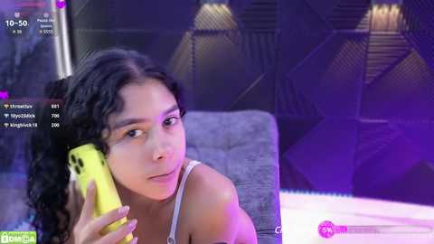 Media: Video of a young woman with curly hair and light skin, wearing a white tank top, holding a yellow phone, smiling against a modern, dark-walled background.