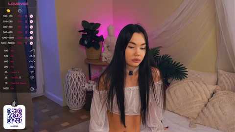 Media: Video of an Asian woman with long black hair, wearing a white off-shoulder crop top, in a cozy bedroom with a green plant, white curtains, and a plush beige blanket.