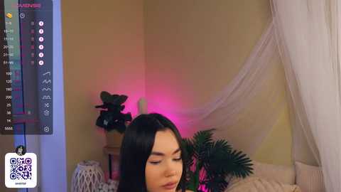 Media: Video of an Asian woman with long black hair and fair skin, sitting on a bed with a white canopy. She has a serene expression. Background features a potted plant and a glowing pink light.