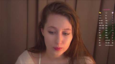 Media: Video of a young Caucasian woman with light skin, long brown hair, and minimal makeup, wearing a white top, gazing downwards against a brown wooden backdrop.