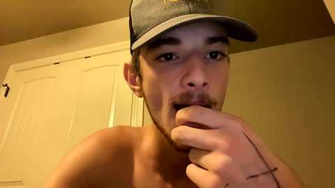 Media: Video of a young, shirtless man with a light complexion, wearing a gray baseball cap, and biting his finger, standing in a beige-walled room with white closet doors in the background.