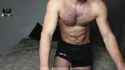 Media: Video of a shirtless, muscular, light-skinned man with a hairy chest, wearing black shorts, sitting on a bed with gray sheets in a dimly lit room.