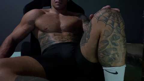 Media: Video of a muscular, tattooed man with a tan, sitting on a black chair, wearing black briefs and white socks, showcasing his muscular physique and intricate body art.