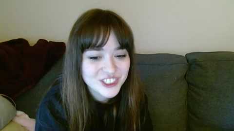 Media: Video of a young Caucasian woman with long brown hair and bangs, wearing a dark shirt, smiling, seated on a gray couch against a beige wall.