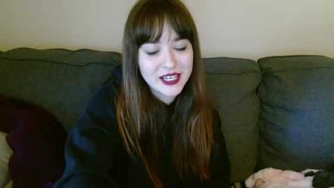 Media: A video of a young woman with long brown hair, wearing dark lipstick, smiling while sitting on a gray couch.