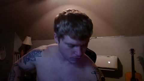 Media: Video of a shirtless man with short, curly brown hair, wearing a black t-shirt, and tattoos on his left shoulder and right arm, in a dimly-lit room with a lamp, air conditioner, and acoustic guitar.