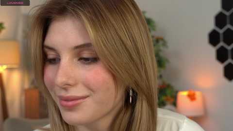 Media: Video of a fair-skinned woman with straight, light brown hair, wearing minimal makeup, smiling softly. Background features a cozy indoor setting with a lamp, hexagonal wall art, and a candlelit table.