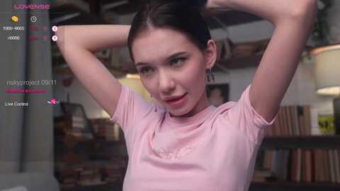 Media: A video of a young woman with fair skin, dark hair, and small breasts, wearing a pink T-shirt, lifting her arms, in a dimly lit room with shelves and a lamp.