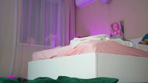 Media: Video of a cluttered, beige bedroom with a white bed, green pillows, and a partially visible person under pink sheets. A framed picture and an air conditioner are on the wall.