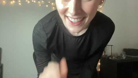 Media: Video of a smiling, fair-skinned young woman with short hair, wearing a black long-sleeve shirt, giving a thumbs-up. Background features a string of warm fairy lights, a desk with a monitor, and a chair.