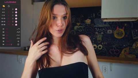 Media: Video of a young woman with long, reddish-brown hair, wearing a black strapless dress, in a kitchen with chalkboard walls adorned with colorful chalk drawings.