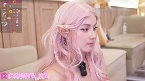 Media: Video of a young woman with long, wavy pink hair, pale skin, and elf ears, wearing a black choker. She sits on a beige sofa in a cozy, well-lit room.