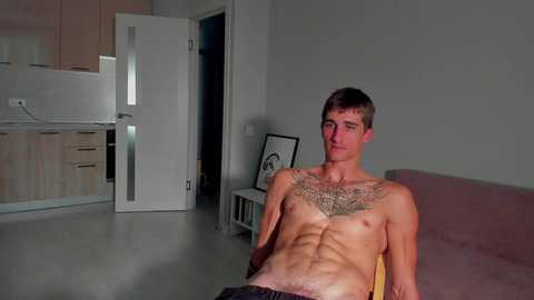 Media: Video of a shirtless young Caucasian man with a lean physique, tattoos on his chest, sitting on a couch in a modern, sparsely furnished room with light wood cabinets and a white door.