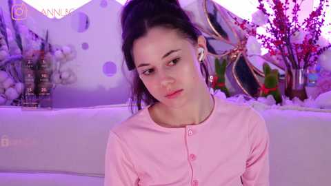 Media: Video of a young woman with pale skin and dark hair in a ponytail, wearing white headphones and a pink blouse, seated on a white couch. Background features a winter-themed mural with snowflakes and trees.